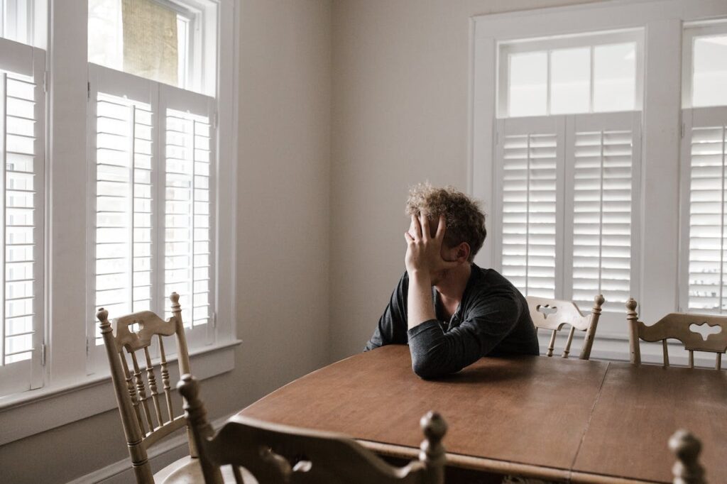 A man cradling his head in his hand sitting alone at a wooden dining table in a barely furnished room with sunlight streaming through plantation shutters
